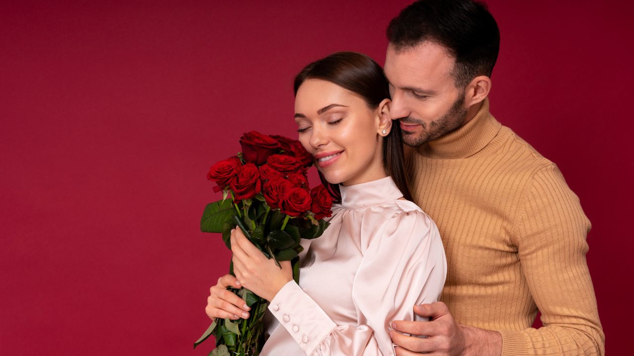 Imagem de um casal a frente de um fundo neutro. A mulher está usando uma camiseta clara e segurando um buquê de flores. O homem está atrás dela, abraçando-a.